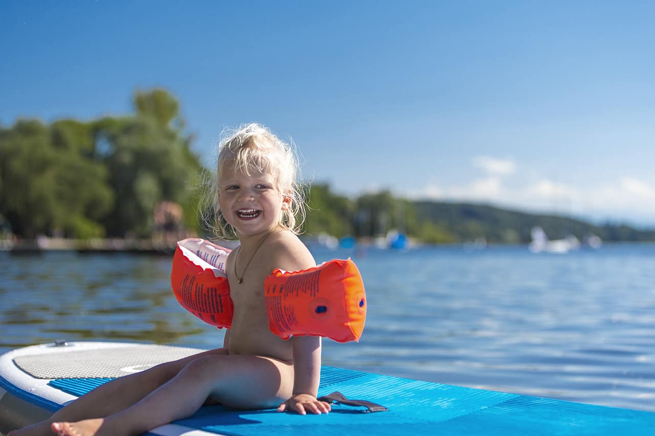 Mädchen mit Schwimmflügeln sitzt auf schwimmendem Stand Up Paddle am Walchensee